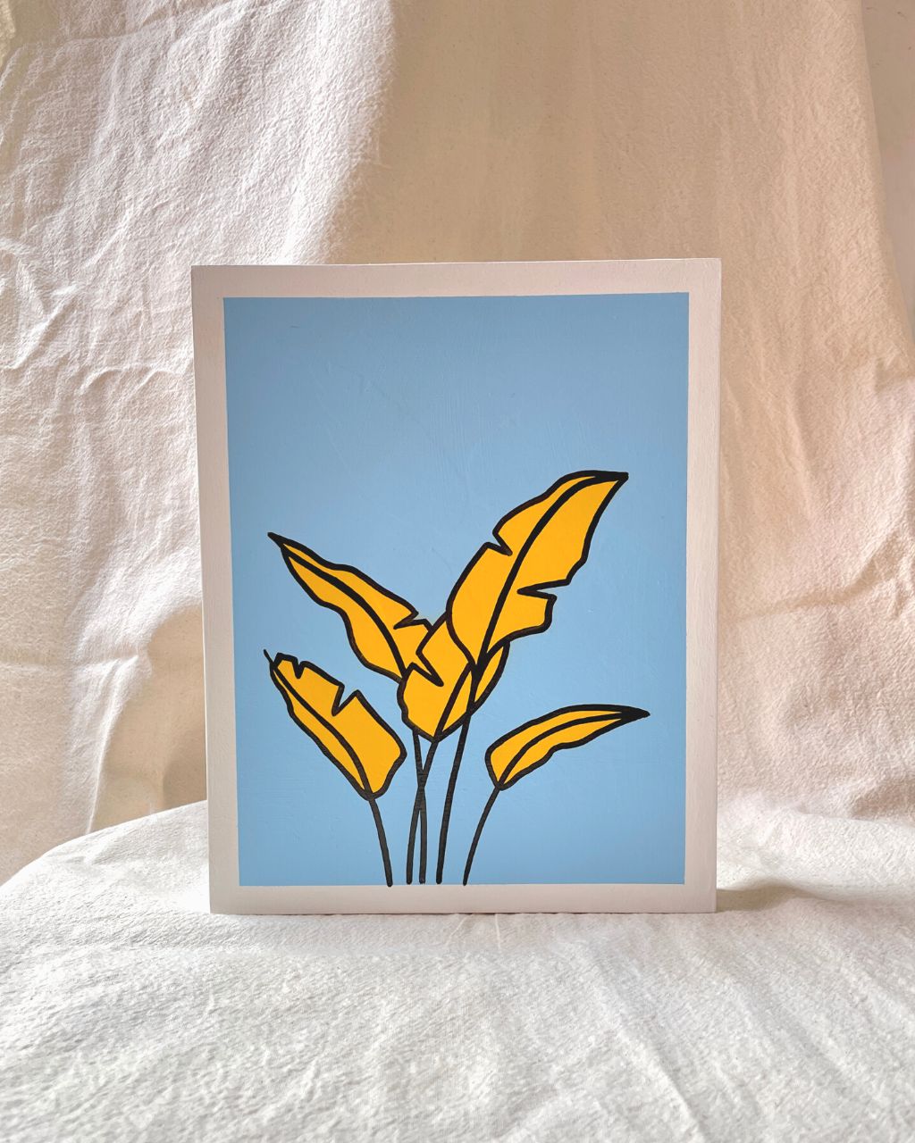 Arte con la técnica de línea clara que ilustra una hoja de plátano amarillo sobre color azul cielo, tamaño 19.5 x 24.5 cm